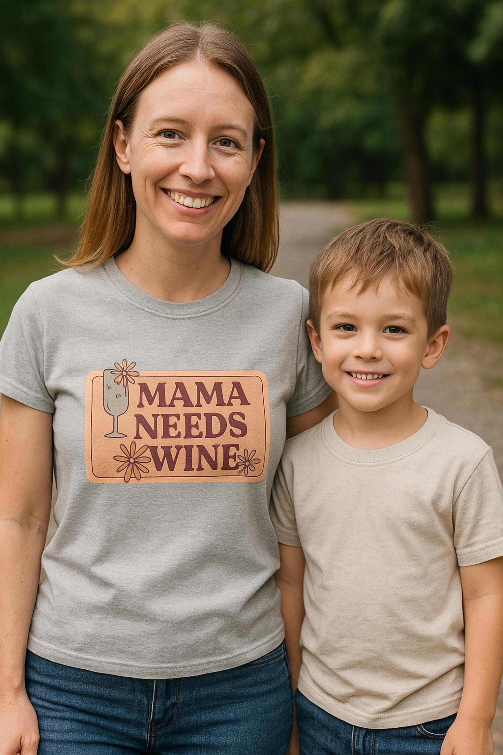 Smiling woman wearing a light gray t-shirt with a playful retro-style graphic that says &quot;Mama Needs Wine&quot; in bold maroon letters on a peach background, accented with a wine glass and flower illustrations. She is standing outdoors on a tree-lined path next to a smiling young boy in a beige t-shirt, both enjoying a casual day in the park.
