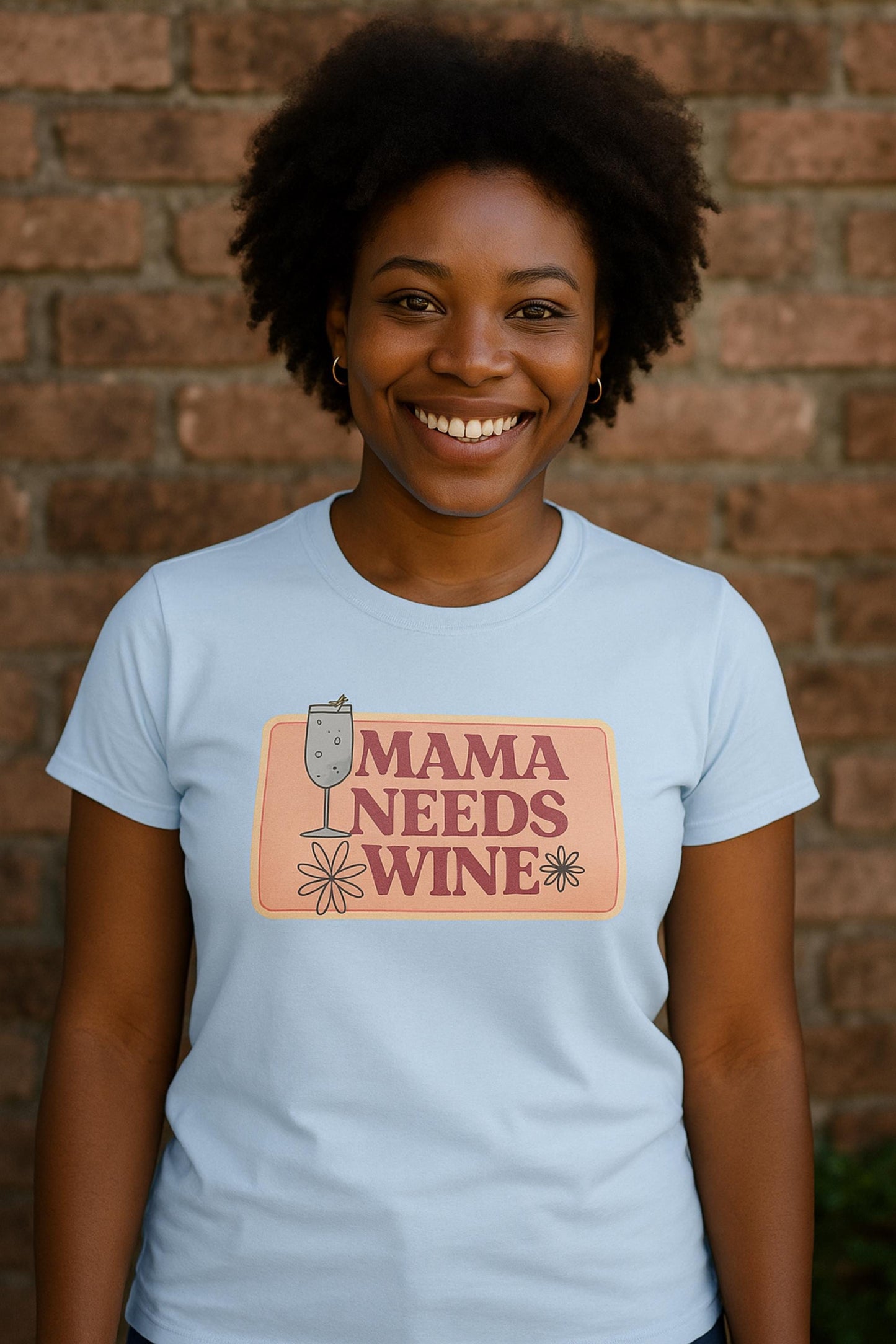 Smiling woman wearing a light blue fitted t-shirt with a retro-style graphic that reads &quot;Mama Needs Wine&quot; in bold pink letters on a peach-colored background. The design features playful daisy flowers and a wine glass illustration. She is standing outdoors in front of a rustic brick wall.