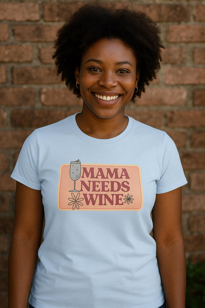 Smiling woman wearing a light blue fitted t-shirt with a retro-style graphic that reads &quot;Mama Needs Wine&quot; in bold pink letters on a peach-colored background. The design features playful daisy flowers and a wine glass illustration. She is standing outdoors in front of a rustic brick wall.