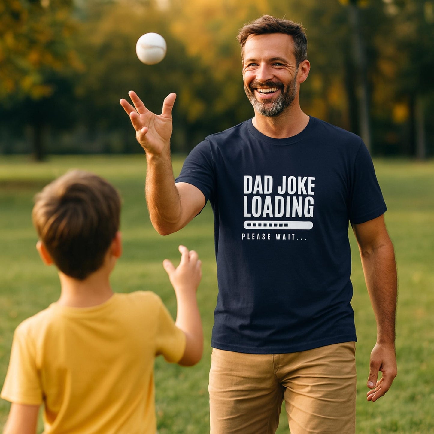 Man wearing a navy blue T-shirt with the text “Dad Joke Loading… Please Wait” and a progress bar graphic in white, smiling while tossing a baseball to a child in a yellow shirt, outdoors in a sunny park.