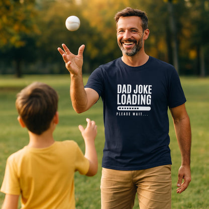 Man wearing a navy blue T-shirt with the text “Dad Joke Loading… Please Wait” and a progress bar graphic in white, smiling while tossing a baseball to a child in a yellow shirt, outdoors in a sunny park.