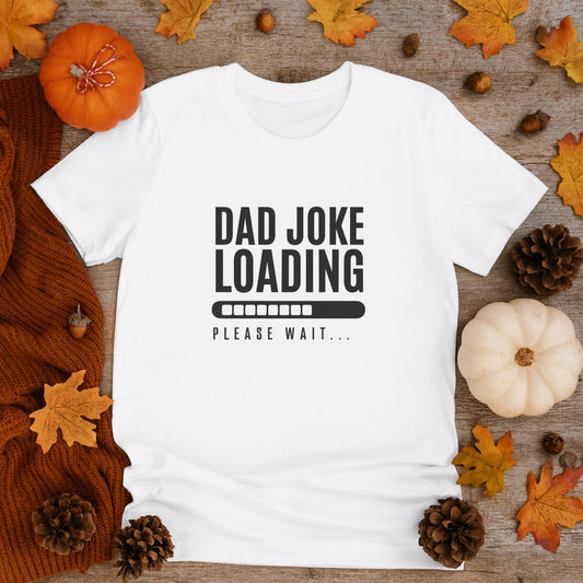White T-shirt with the text “Dad Joke Loading… Please Wait” and a progress bar graphic in black, displayed flat on a wooden surface surrounded by autumn decorations including pumpkins, pinecones, acorns, orange knit fabric, and colorful fall leaves.