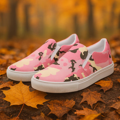 Pair of slip-on canvas shoes with a pink, cream, and black camouflage pattern, featuring white rubber soles, displayed outdoors on autumn leaves with a blurred forest background.