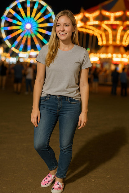 Woman standing at a nighttime carnival wearing a light gray t-shirt, blue jeans, and pink camouflage slip-on shoes, with a brightly lit Ferris wheel and carousel glowing in the blurred background.