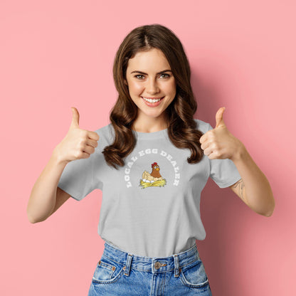Smiling woman giving two thumbs up while wearing a light gray t-shirt with a cute illustration of a chicken sitting on a nest. The shirt features the phrase &quot;LOCAL EGG DEALER&quot; in bold white letters. She stands in front of a solid pink background, exuding a cheerful and playful vibe.