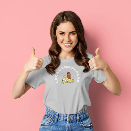 Smiling woman giving two thumbs up while wearing a light gray t-shirt with a cute illustration of a chicken sitting on a nest. The shirt features the phrase &quot;LOCAL EGG DEALER&quot; in bold white letters. She stands in front of a solid pink background, exuding a cheerful and playful vibe.
