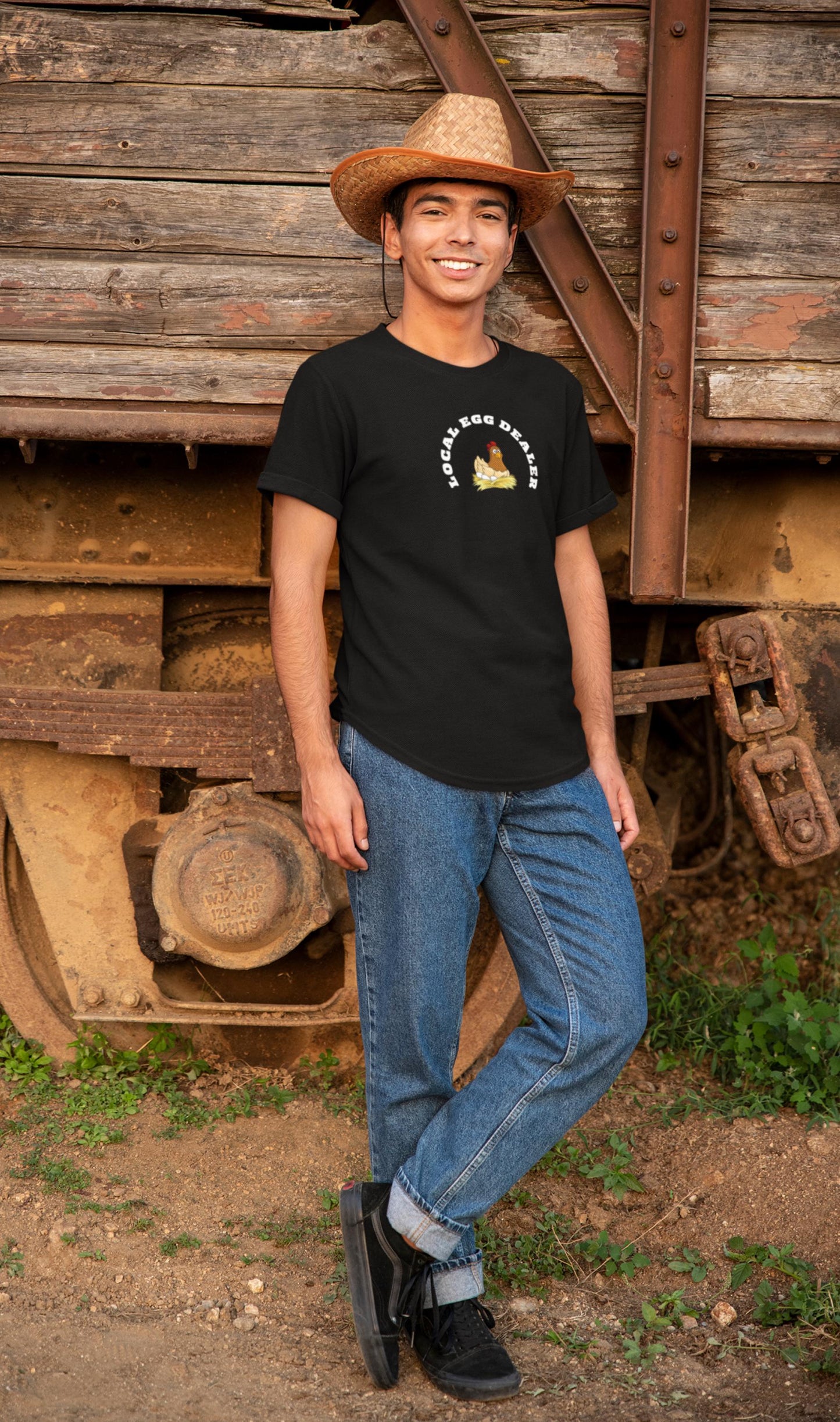 Smiling young man wearing a straw cowboy hat, black t-shirt with a cartoon hen on a nest and the phrase &quot;LOCAL EGG DEALER&quot; in white text. He stands casually against a rustic wooden and metal train car, dressed in cuffed blue jeans and black sneakers, with a dirt ground and greenery around him.