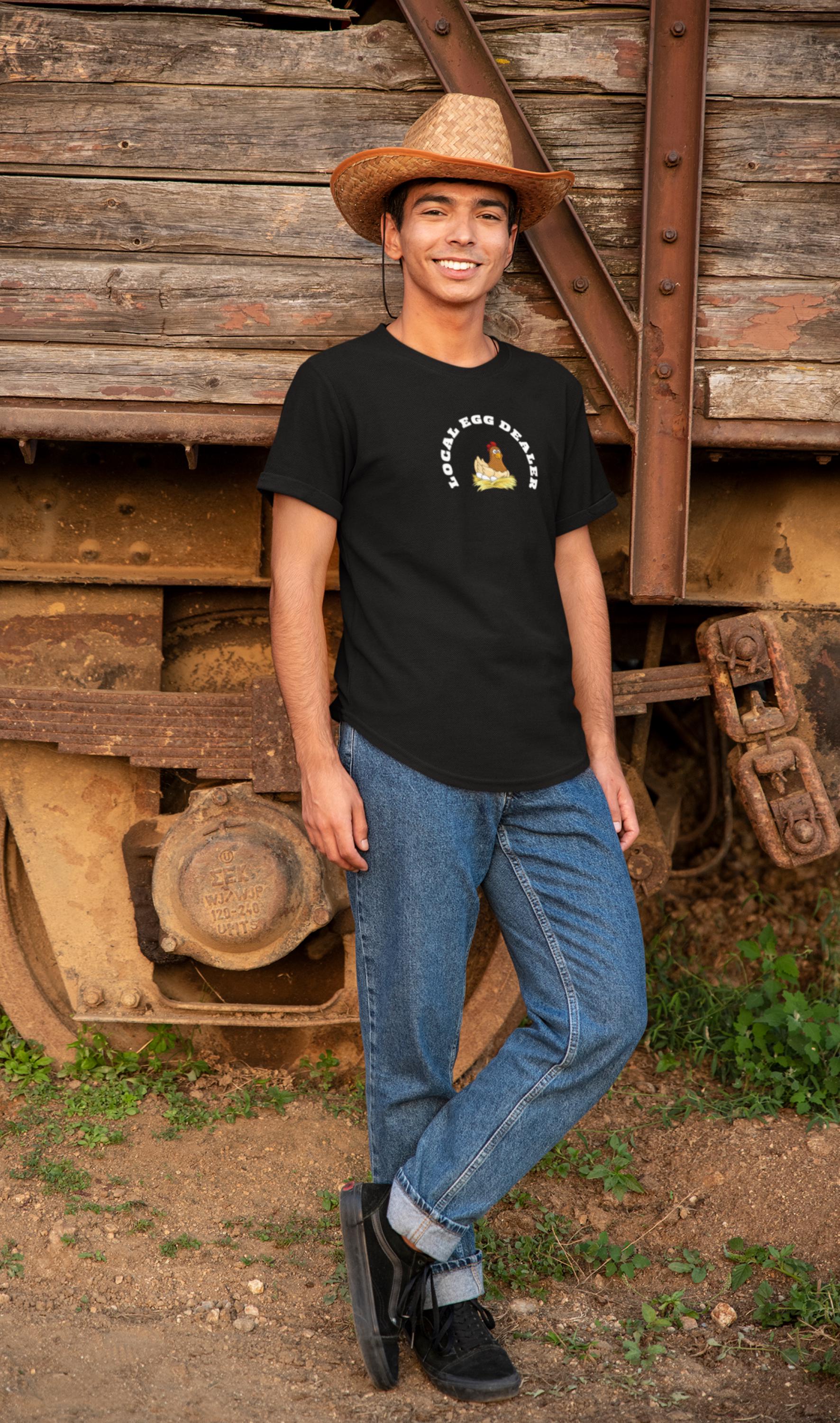 Smiling young man wearing a straw cowboy hat, black t-shirt with a cartoon hen on a nest and the phrase &quot;LOCAL EGG DEALER&quot; in white text. He stands casually against a rustic wooden and metal train car, dressed in cuffed blue jeans and black sneakers, with a dirt ground and greenery around him.