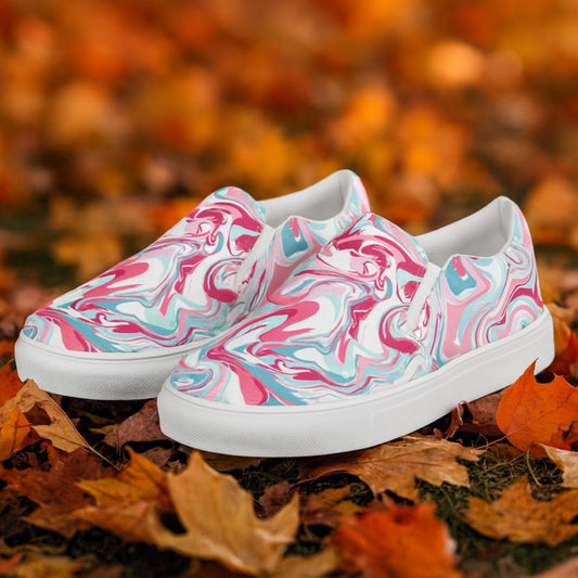 A pair of slip-on canvas sneakers with a vibrant pink, white, and turquoise marbled swirl pattern, displayed outdoors on a ground covered with colorful autumn leaves.