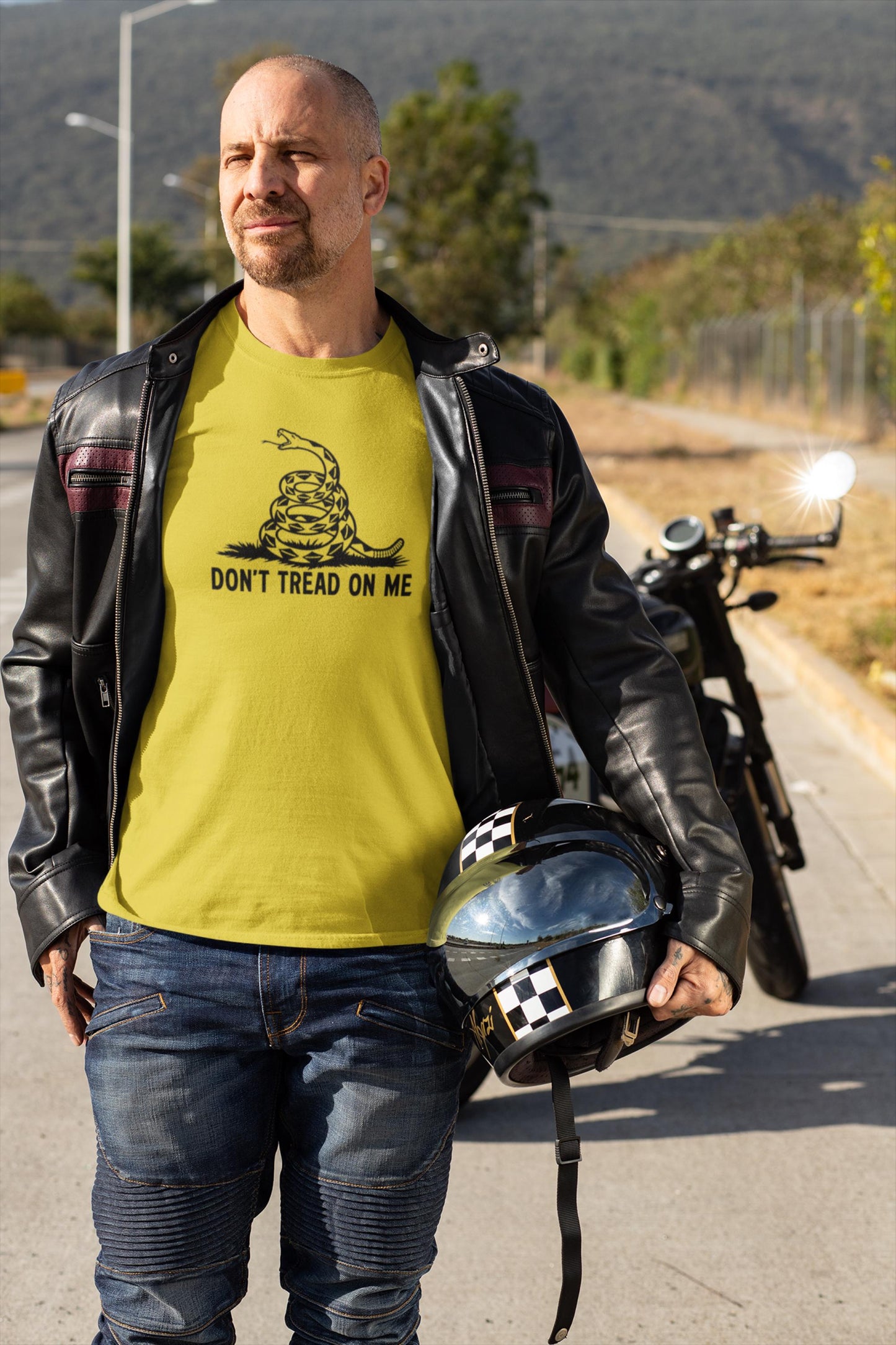 Man standing outdoors beside a motorcycle, wearing a yellow &quot;Don&#39;t Tread On Me&quot; t-shirt with a bold black coiled rattlesnake graphic. He has a black leather jacket draped over his shoulders and is holding a black helmet with checkered details. The scene is sunlit with mountains and trees in the background, suggesting a scenic ride.