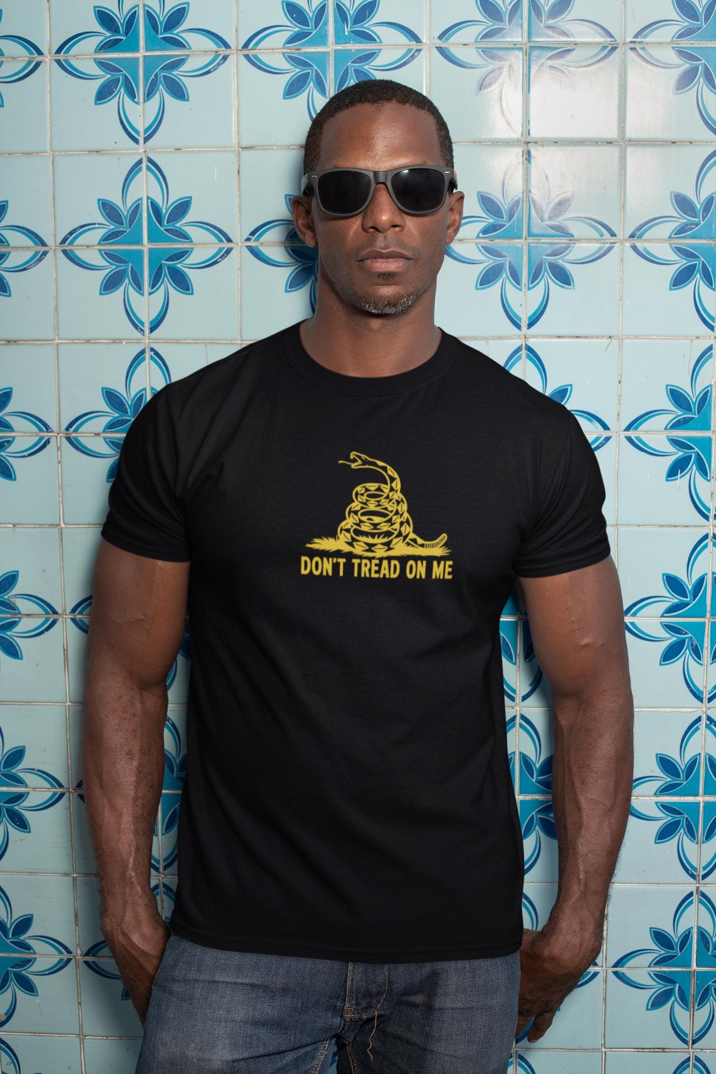 Man wearing sunglasses and a bright yellow &quot;Don&#39;t Tread On Me&quot; t-shirt featuring a black coiled rattlesnake graphic. He stands confidently in front of a decorative blue and white tiled wall with floral patterns, exuding a bold and assertive vibe.