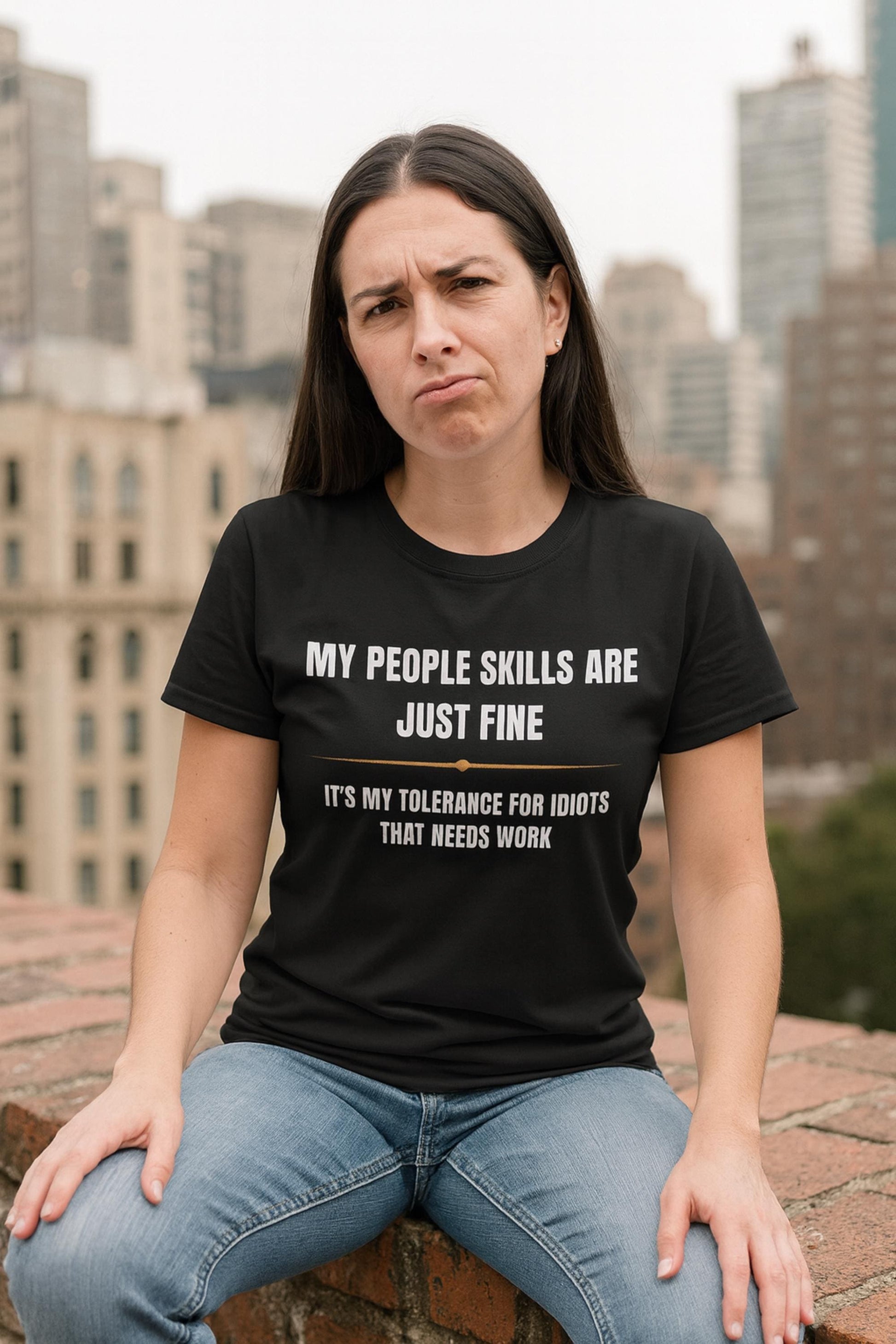 Woman wearing a black T-shirt with bold white and gold text that reads, “MY PEOPLE SKILLS ARE JUST FINE — IT’S MY TOLERANCE FOR IDIOTS THAT NEEDS WORK.” She is sitting on a rooftop ledge with a skeptical or unimpressed facial expression, set against a backdrop of city buildings.