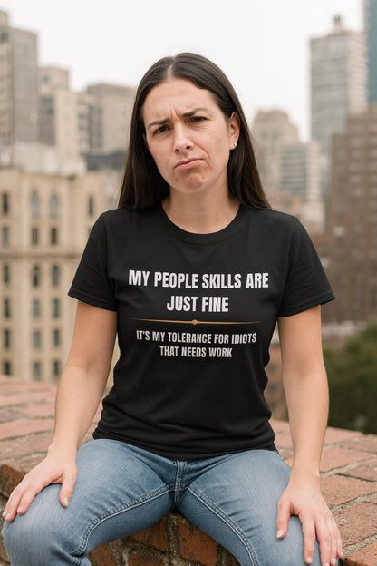 Woman wearing a black T-shirt with bold white and gold text that reads, “MY PEOPLE SKILLS ARE JUST FINE — IT’S MY TOLERANCE FOR IDIOTS THAT NEEDS WORK.” She is sitting on a rooftop ledge with a skeptical or unimpressed facial expression, set against a backdrop of city buildings.