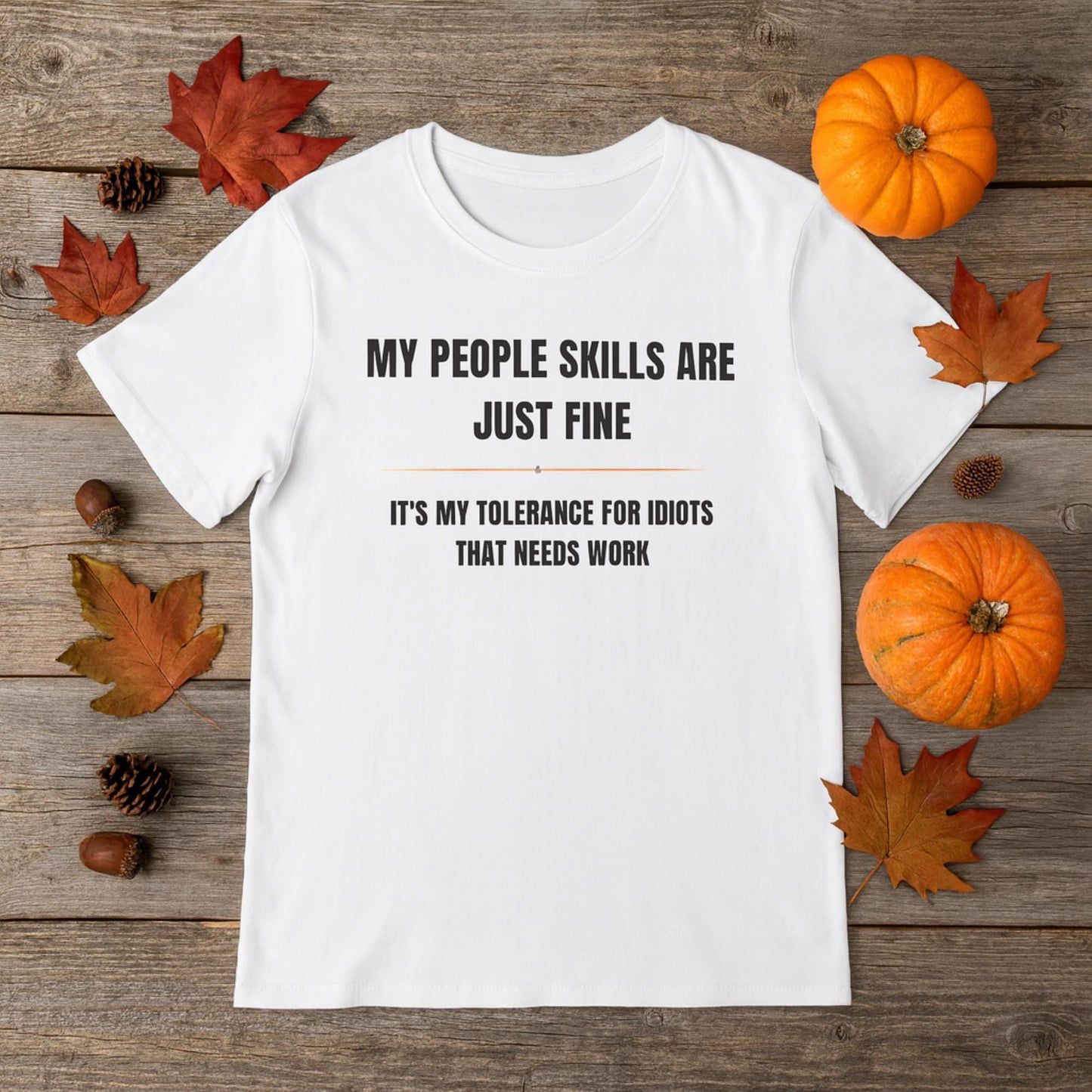 Flat lay of a white T-shirt with bold black text that reads, “MY PEOPLE SKILLS ARE JUST FINE. IT’S MY TOLERANCE FOR IDIOTS THAT NEEDS WORK,” styled with autumn-themed décor including pumpkins, acorns, pinecones, and colorful fall leaves on a rustic wooden background.