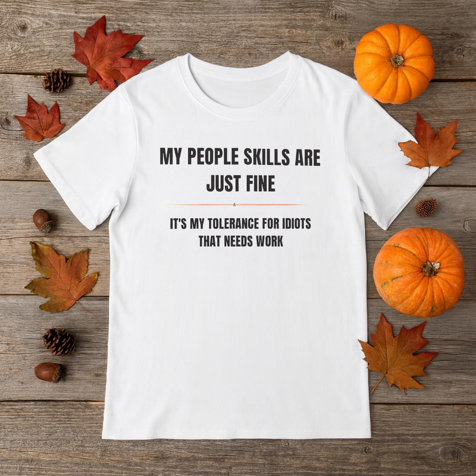 Flat lay of a white T-shirt with bold black text that reads, “MY PEOPLE SKILLS ARE JUST FINE. IT’S MY TOLERANCE FOR IDIOTS THAT NEEDS WORK,” styled with autumn-themed décor including pumpkins, acorns, pinecones, and colorful fall leaves on a rustic wooden background.