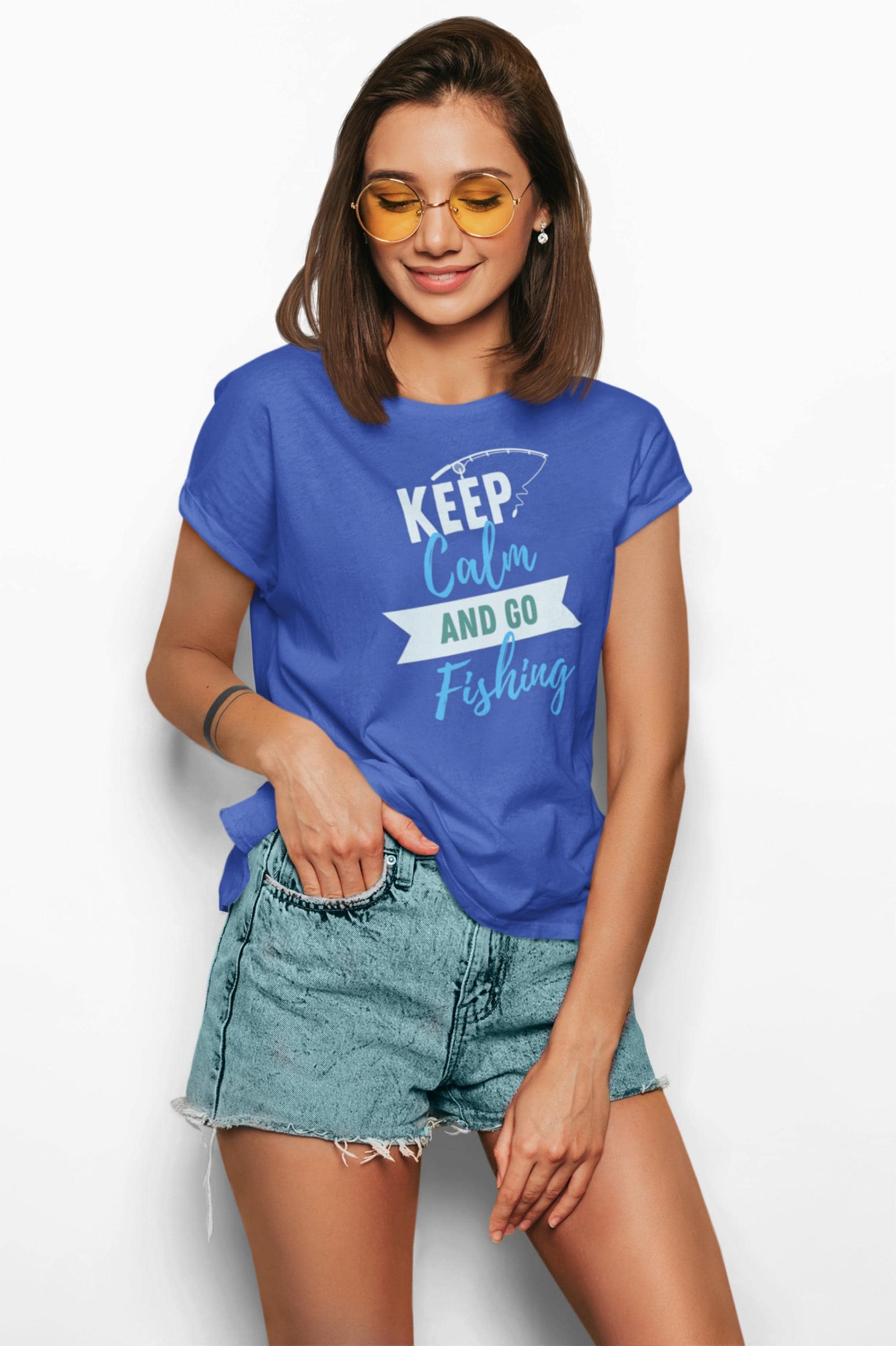Smiling young woman wearing yellow-tinted sunglasses and a royal blue T-shirt with a playful fishing-themed graphic. The shirt reads “KEEP Calm AND GO Fishing” in white, teal, and light blue text with a fishing hook illustration. She pairs the shirt with light denim high-waisted shorts and poses casually against a white background.