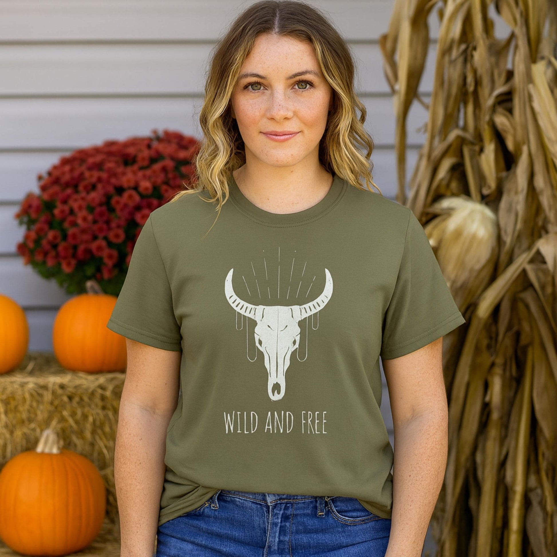 Woman standing outdoors in a fall setting with pumpkins, hay bales, and dried corn stalks. She is wearing an olive green T-shirt featuring a graphic of a bull skull with decorative lines above it and the text “WILD AND FREE” below. She pairs the shirt with blue jeans and has wavy hair.
