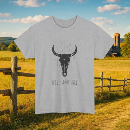 Light gray T-shirt hanging outdoors against a rustic countryside background with a wooden fence, open field, and barn in the distance. The shirt features a large graphic of a black bull skull with decorative lines above it and the text “WILD AND FREE” below.