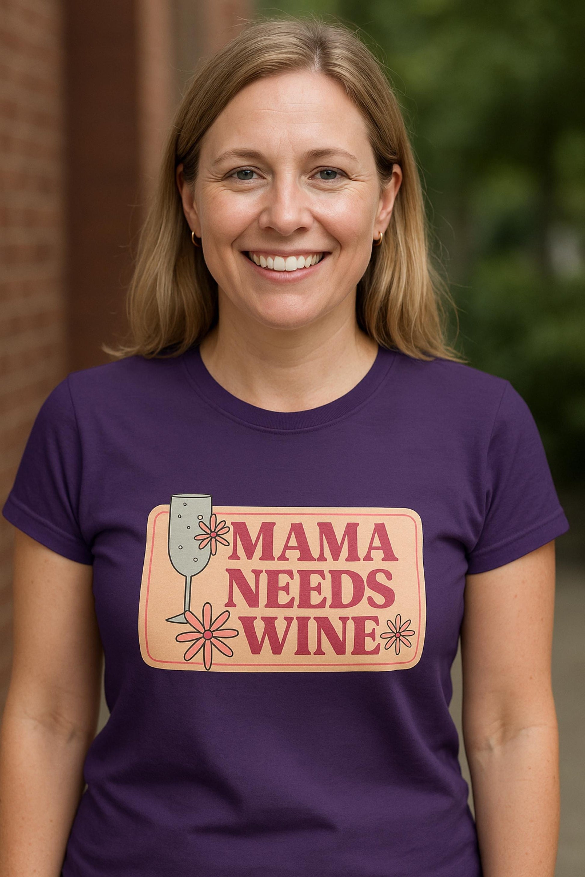 Smiling woman wearing a fitted purple t-shirt with a playful retro-style graphic that says &quot;Mama Needs Wine&quot; in bold pink lettering on a peach background. The design features a wine glass and whimsical flower accents. She is standing outside with a brick wall and greenery in the background.
