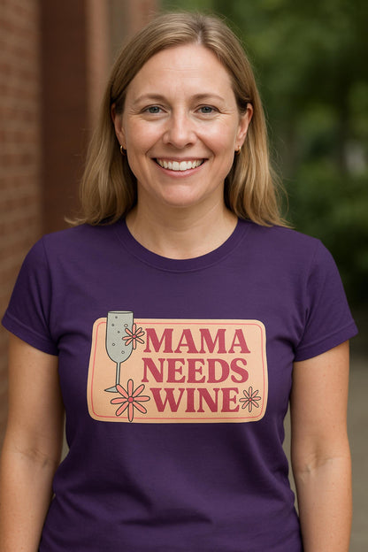 Smiling woman wearing a fitted purple t-shirt with a playful retro-style graphic that says &quot;Mama Needs Wine&quot; in bold pink lettering on a peach background. The design features a wine glass and whimsical flower accents. She is standing outside with a brick wall and greenery in the background.
