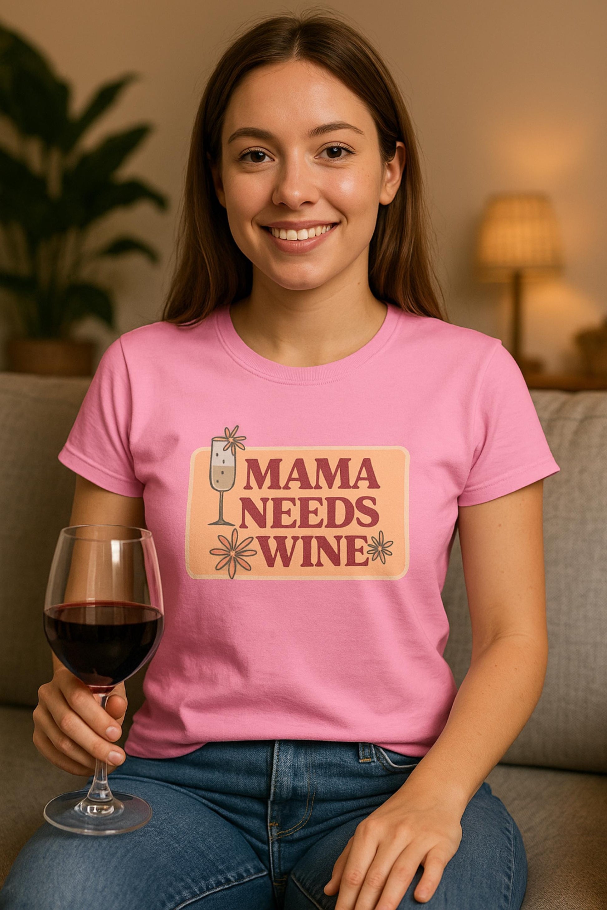 Smiling woman sitting on a couch wearing a pink t-shirt with a retro-style graphic that says &quot;Mama Needs Wine&quot; in bold maroon text on a peach background. The design includes a champagne glass and playful flower accents. She is holding a glass of red wine, with a cozy lamp and plant in the softly lit background.