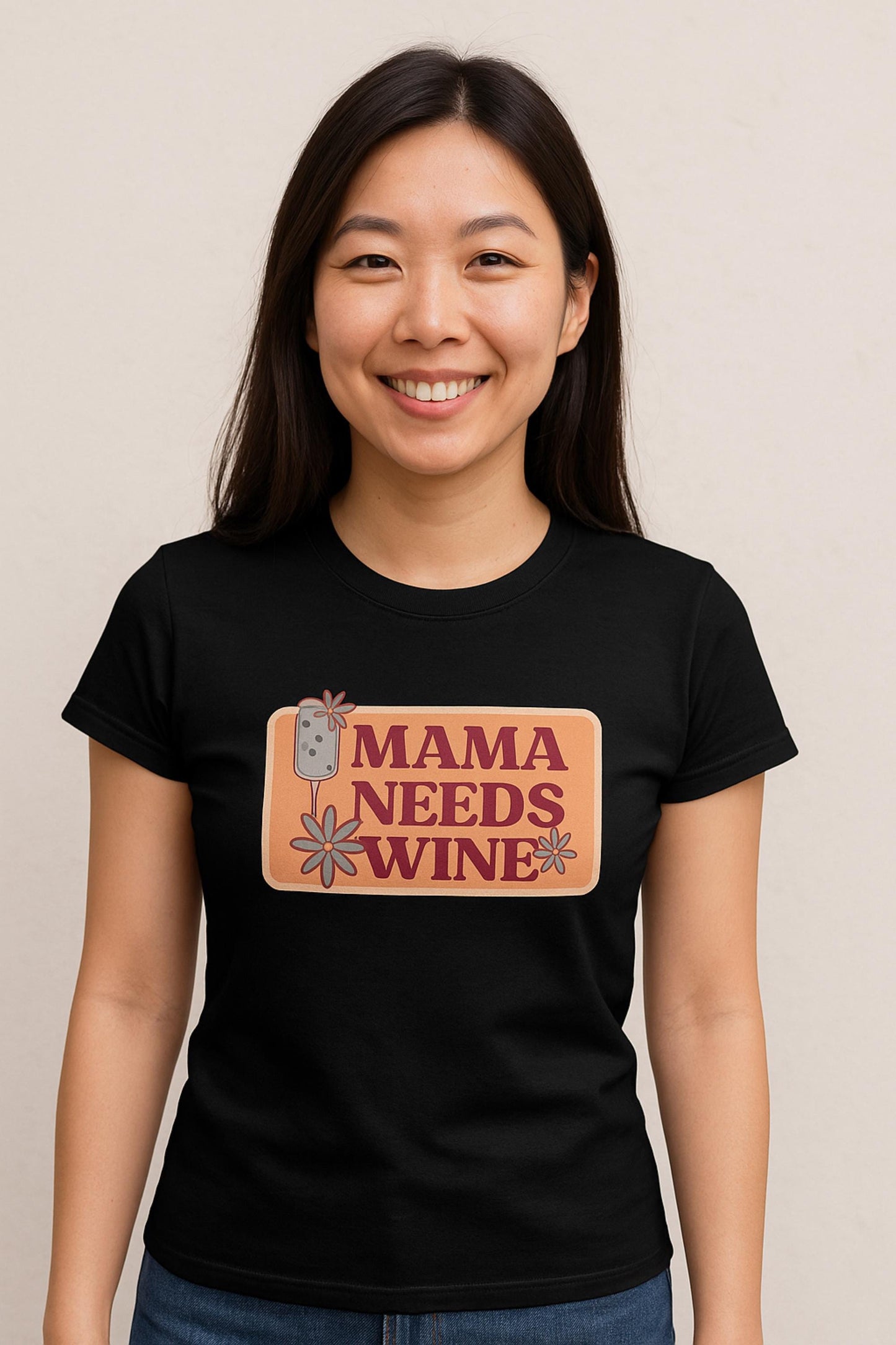 Smiling woman wearing a fitted black t-shirt with a retro-style graphic that says &quot;Mama Needs Wine&quot; in bold burgundy letters on a peach-colored background. The design includes a wine glass and whimsical daisy flowers, giving it a fun and playful vibe. She stands in front of a neutral light beige background.