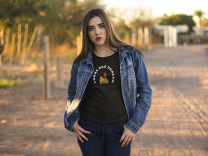 Woman standing outdoors on a dirt path during golden hour, wearing a black t-shirt with a cartoon hen sitting on a nest and the phrase &quot;LOCAL EGG DEALER&quot; in white text. She has a serious expression, layered with a denim jacket and jeans, with trees and a rural setting in the background.