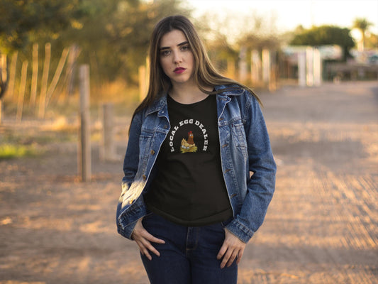 Woman standing outdoors on a dirt path during golden hour, wearing a black t-shirt with a cartoon hen sitting on a nest and the phrase &quot;LOCAL EGG DEALER&quot; in white text. She has a serious expression, layered with a denim jacket and jeans, with trees and a rural setting in the background.
