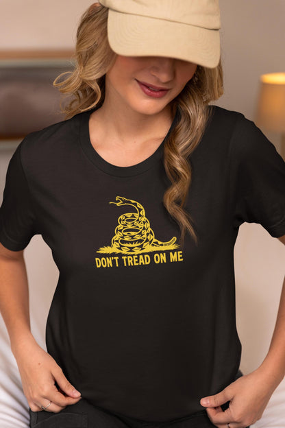 Woman wearing a black t-shirt with a bold yellow &quot;Don&#39;t Tread On Me&quot; graphic featuring a coiled rattlesnake, inspired by the Gadsden flag. She is sitting indoors, slightly looking down with a soft smile, wearing a beige baseball cap. The setting appears cozy and warmly lit.