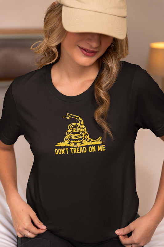 Woman wearing a black t-shirt with a bold yellow &quot;Don&#39;t Tread On Me&quot; graphic featuring a coiled rattlesnake, inspired by the Gadsden flag. She is sitting indoors, slightly looking down with a soft smile, wearing a beige baseball cap. The setting appears cozy and warmly lit.
