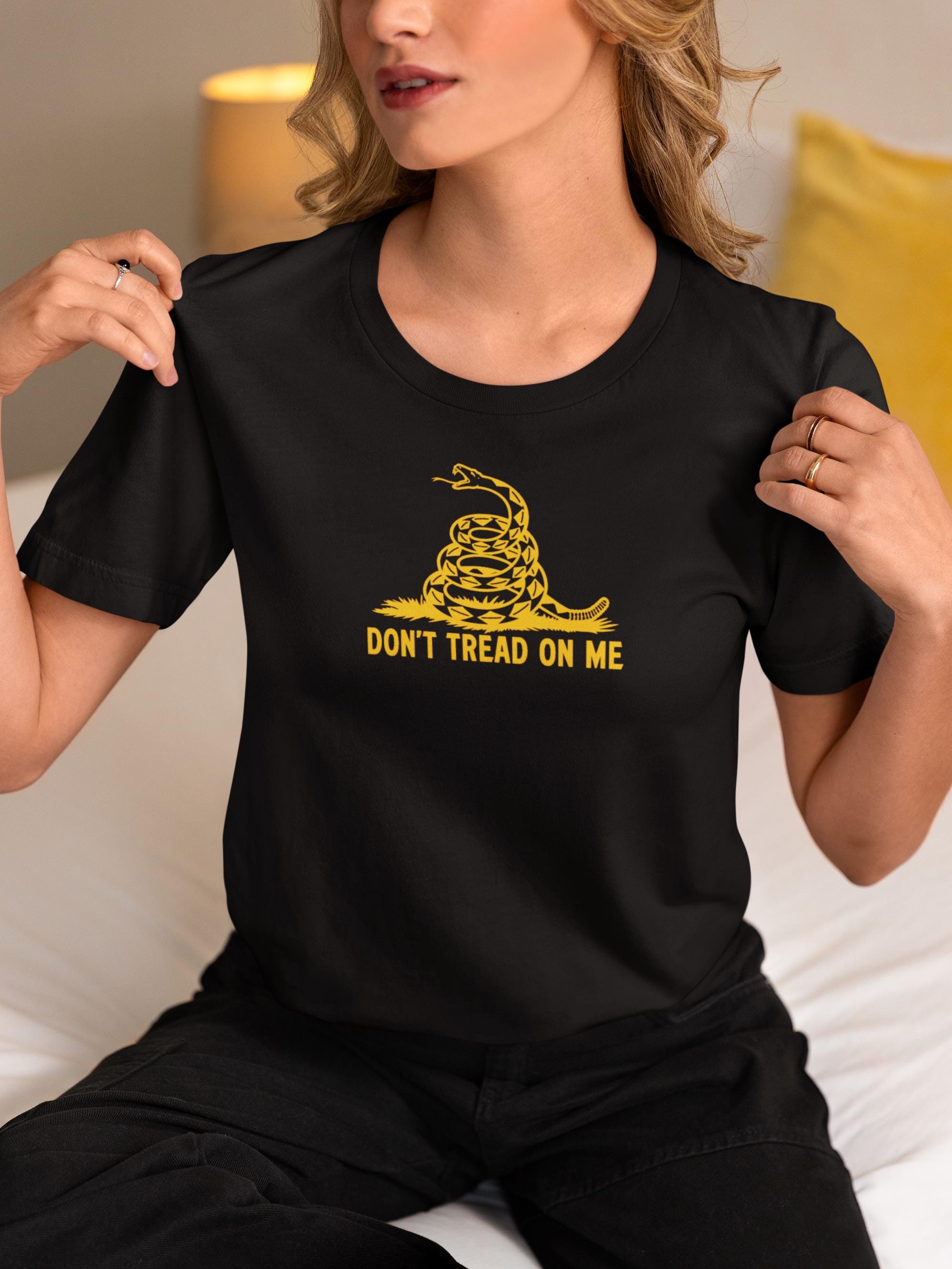 Woman sitting indoors on a bed wearing a black t-shirt featuring a bold yellow &quot;Don&#39;t Tread On Me&quot; graphic with a coiled rattlesnake, inspired by the Gadsden flag. She is pulling slightly at the collar of the shirt, showcasing the design, with a softly lit background and a yellow pillow behind her.