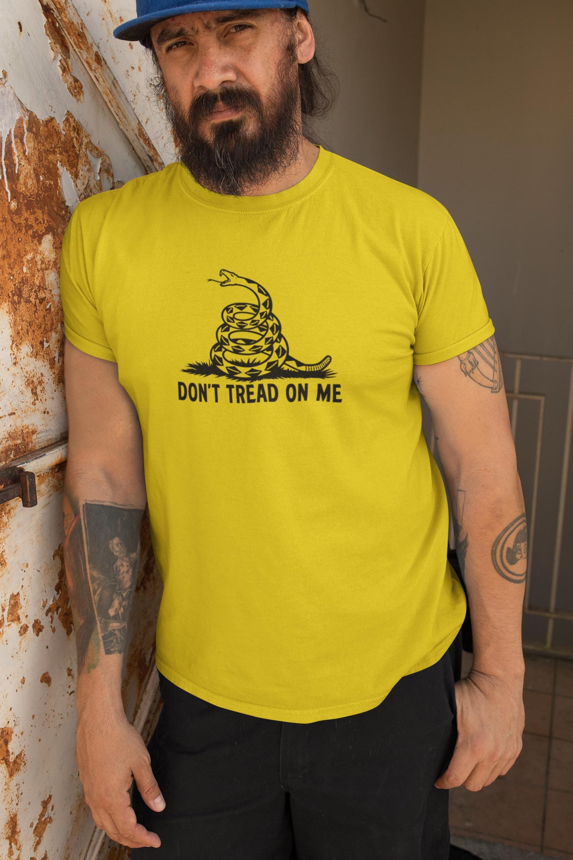 Man with a beard and tattoos wearing a bright yellow &quot;Don&#39;t Tread On Me&quot; t-shirt featuring a black coiled rattlesnake design. He stands against a weathered, rusted metal surface, adding a gritty, rugged backdrop that complements his serious expression and casual streetwear style.