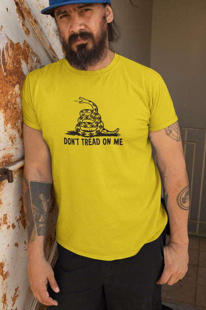 Man with a beard and tattoos wearing a bright yellow &quot;Don&#39;t Tread On Me&quot; t-shirt featuring a black coiled rattlesnake design. He stands against a weathered, rusted metal surface, adding a gritty, rugged backdrop that complements his serious expression and casual streetwear style.