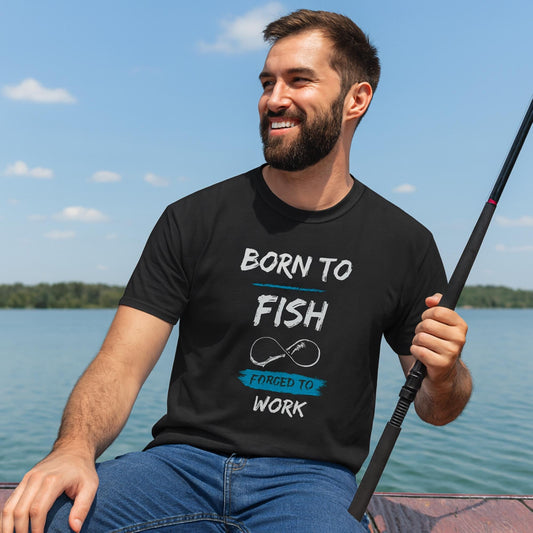 Smiling man sitting outdoors by the water, holding a fishing rod. He is wearing a black T-shirt with the text “BORN TO FISH, FORCED TO WORK” and a graphic of a fishing hook in the shape of an infinity symbol. The background shows a lake under a clear blue sky with a few clouds.