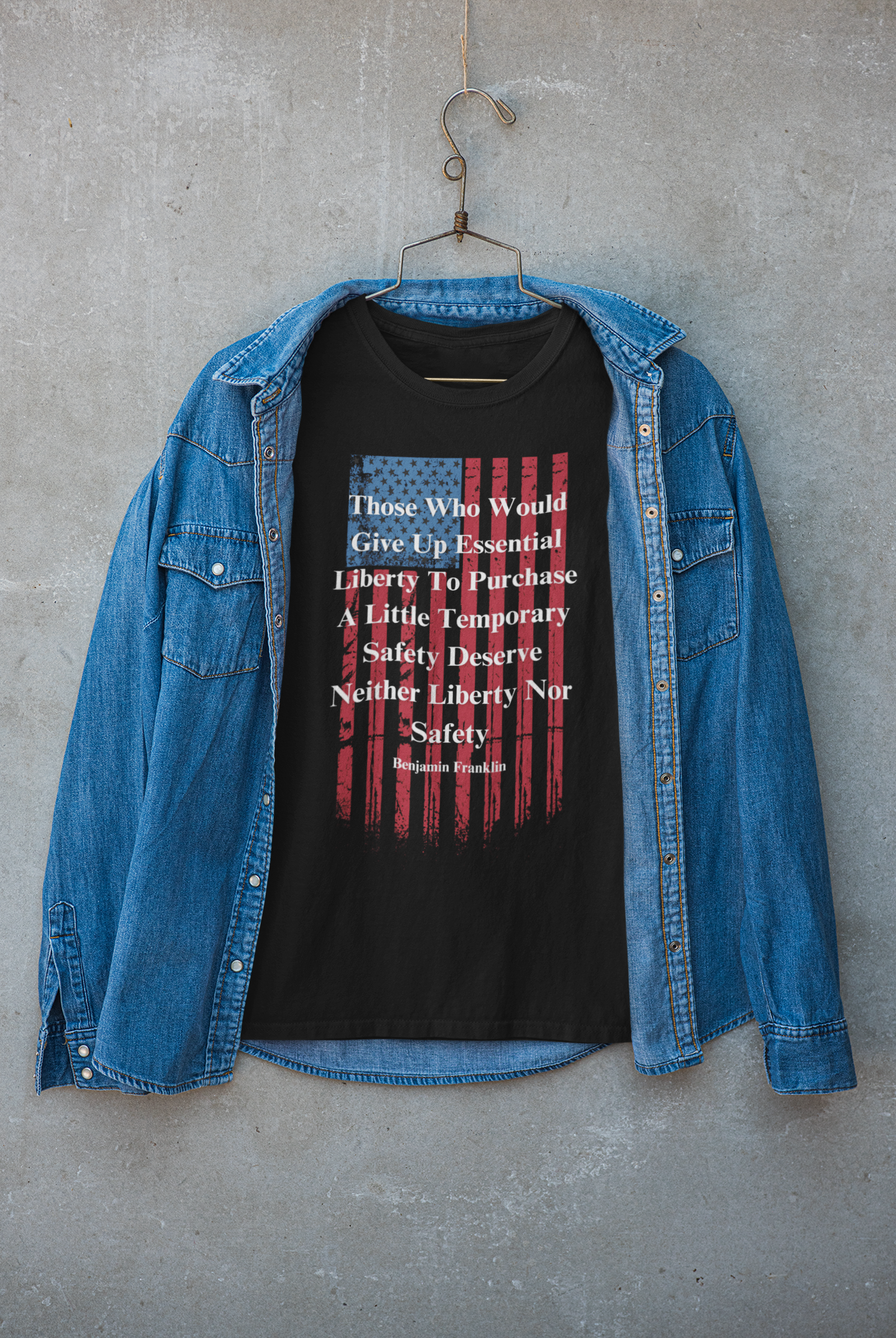 A black T-shirt with a distressed American flag graphic and a bold quote by Benjamin Franklin hangs on a rustic hanger against a concrete wall. The shirt reads: “Those who would give up essential liberty to purchase a little temporary safety deserve neither liberty nor safety.” A denim button-up shirt is draped around the T-shirt, giving the display a casual, rugged, Americana vibe perfect for patriotic or freedom-themed fashion.