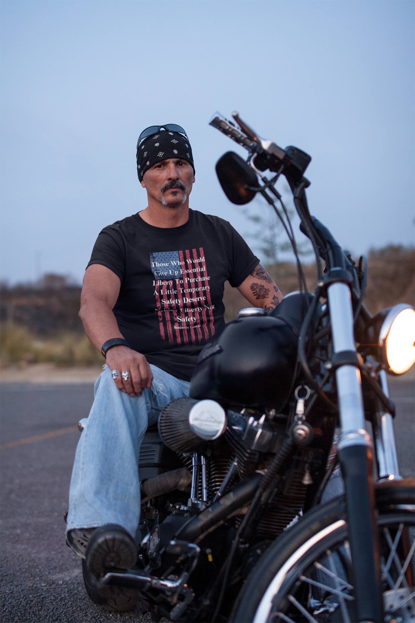 A rugged man with a mustache and bandana sits confidently on a black motorcycle in an outdoor setting at dusk. He wears a black graphic T-shirt featuring a distressed American flag and a quote by Benjamin Franklin: “Those who would give up essential liberty to purchase a little temporary safety deserve neither liberty nor safety.” His look is completed with light blue jeans, rings, a wristband, and sunglasses perched on his head, projecting a tough, patriotic biker aesthetic.