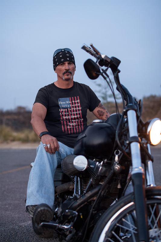 A rugged man with a mustache and bandana sits confidently on a black motorcycle in an outdoor setting at dusk. He wears a black graphic T-shirt featuring a distressed American flag and a quote by Benjamin Franklin: “Those who would give up essential liberty to purchase a little temporary safety deserve neither liberty nor safety.” His look is completed with light blue jeans, rings, a wristband, and sunglasses perched on his head, projecting a tough, patriotic biker aesthetic.
