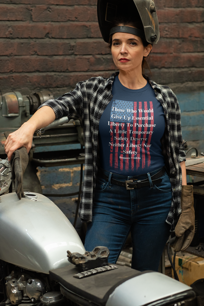 A confident woman wearing a welding helmet lifted on her head stands beside a motorcycle in a garage or workshop. She is dressed in a black-and-white plaid shirt over a navy graphic T-shirt with a distressed American flag and a quote by Benjamin Franklin: “Those who would give up essential liberty to purchase a little temporary safety deserve neither liberty nor safety.” She wears jeans, gloves, and exudes a strong, determined expression, reflecting a patriotic and rugged aesthetic.