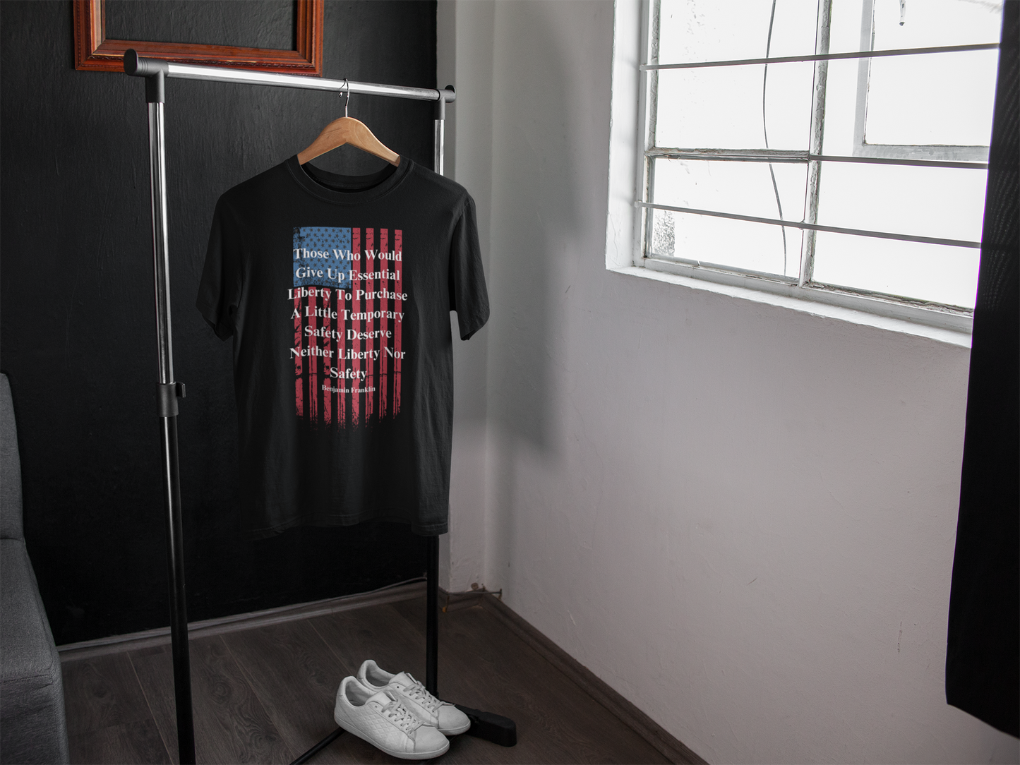 A black T-shirt featuring a distressed American flag graphic and a quote by Benjamin Franklin hangs on a wooden hanger from a minimalist clothing rack in a modern, well-lit room. The quote reads: “Those who would give up essential liberty to purchase a little temporary safety deserve neither liberty nor safety.” A pair of white sneakers rests neatly on the floor beneath the shirt, and natural light streams in from a nearby window, casting soft shadows across the wooden floor and white walls.